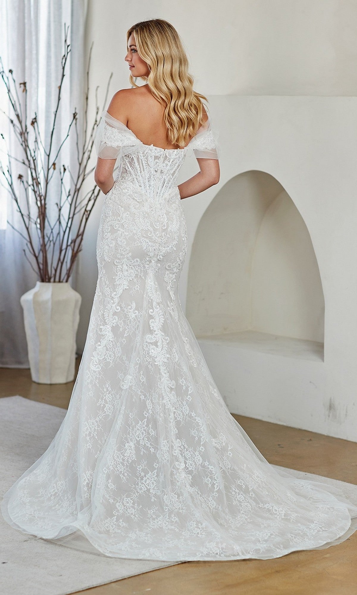 A woman in an off-the-shoulder, lace wedding gown with a fitted bodice and long train, reminiscent of the elegant Long Prom Dress: Amelia Couture 2153, stands indoors facing away from the camera.